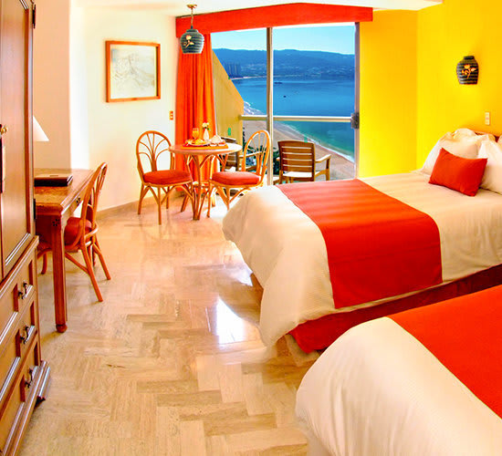 A brightly decorated guest room with two double beds at the Copacabana Beach Acapulco Hotel, featuring vibrant orange accents, tile flooring, and a small table for dining. A large window and balcony offer a stunning, panoramic view of Acapulco Bay. This room setup is ideal for MICE delegates sharing accommodation, offering a comfortable and scenic option for large incentive groups and conference attendees.