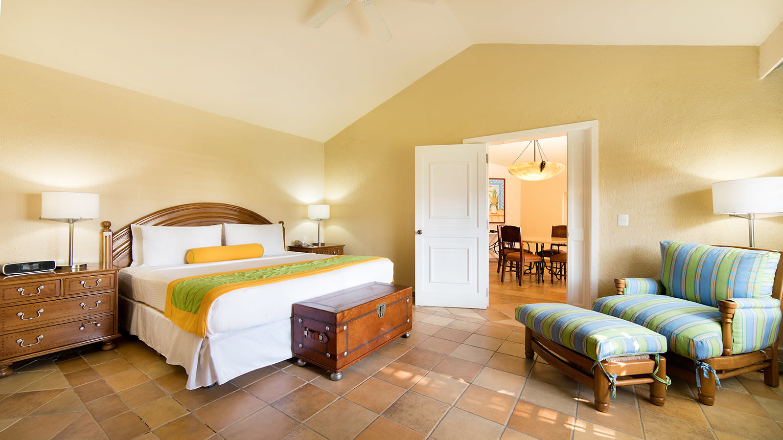 A spacious king bedroom suite at The Pierre Mundo Imperial resort in Acapulco, featuring warm yellow walls, terracotta-colored tile floors, and a dark wooden headboard and furniture. The room opens to a separate living or dining area, confirming its status as a suite. This high-quality accommodation is perfect for VIP MICE delegates, executive retreats, or as a superior room for incentive travel winners, providing comfort and space for business travelers.