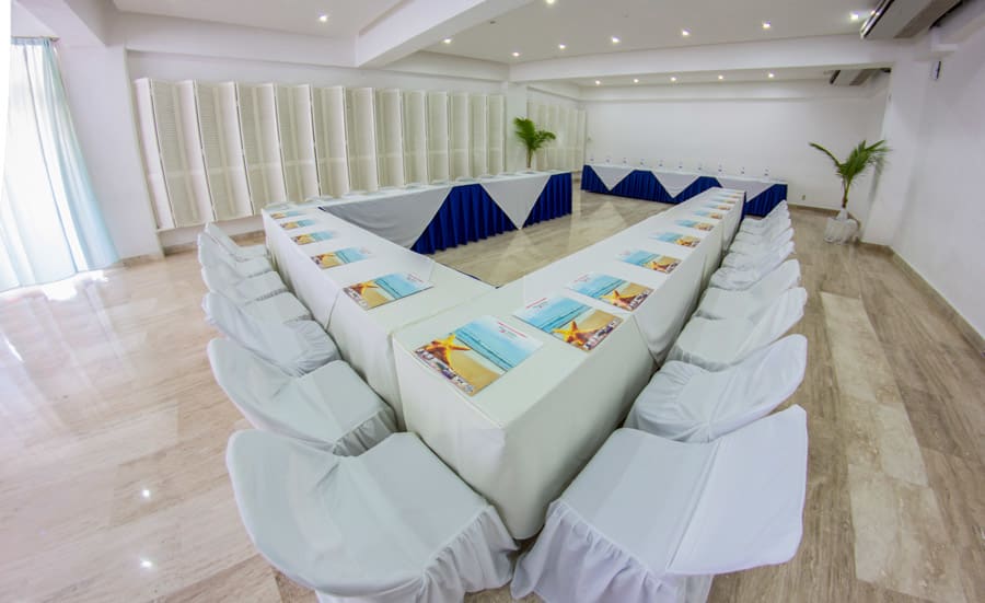A bright, modern meeting room at the Playa Suites Hotel in Acapulco, configured in a U-Shape style for group interaction. The room features a white color palette, marble-like flooring, and white-skirted chairs and tables, providing an elegant and professional environment. This setup is ideal for focused board meetings, interactive training sessions, and high-level MICE discussions.