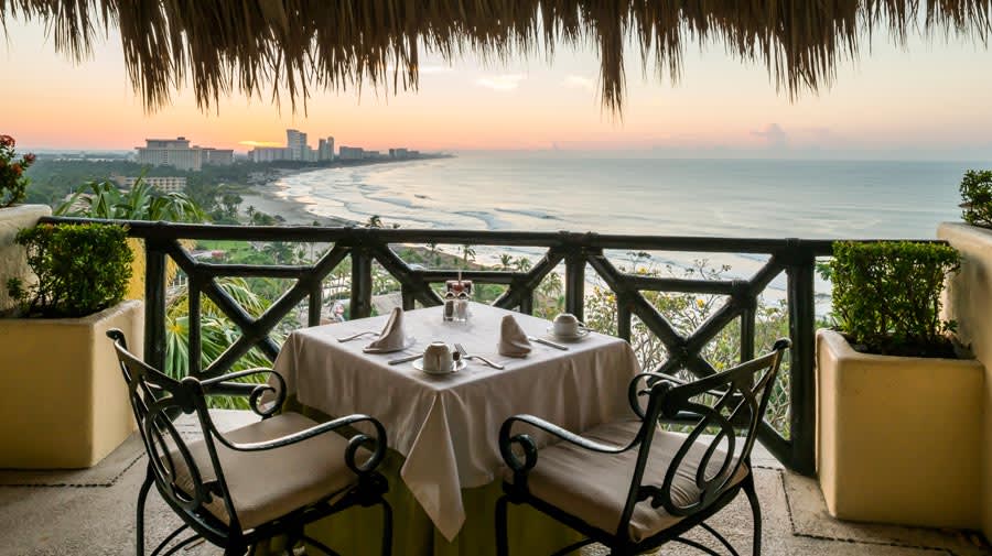 A close-up view of an elegantly set table for two on a private balcony at the Quinta Real Acapulco, overlooking the sprawling bay and beach at sunset. The table setting, featuring white linen and glassware, is framed by a rustic railing and a palapa-style thatched roof. This image highlights the premium, gourmet dining and exclusive corporate hospitality experiences available for incentive travel groups and VIP MICE delegates.