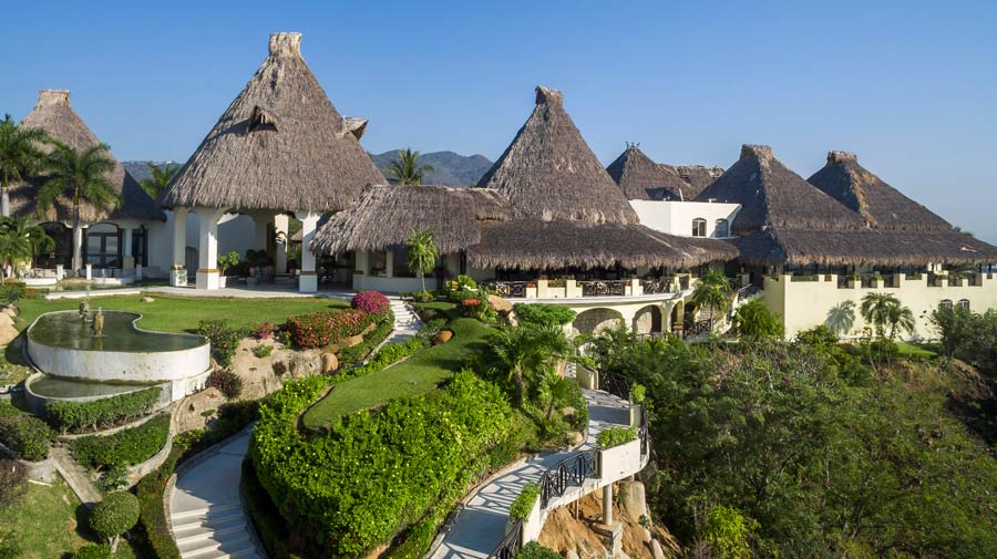 An aerial view of the Quinta Real Acapulco hotel complex, featuring multiple buildings with distinctive, large thatched palapa roofs set amidst manicured gardens and cascading pathways. A tiered, circular fountain is visible on the lawn in the foreground. This unique and picturesque setting highlights the resort's appeal as an exclusive MICE venue for high-end incentive travel, sophisticated corporate retreats, and unique outdoor event space.