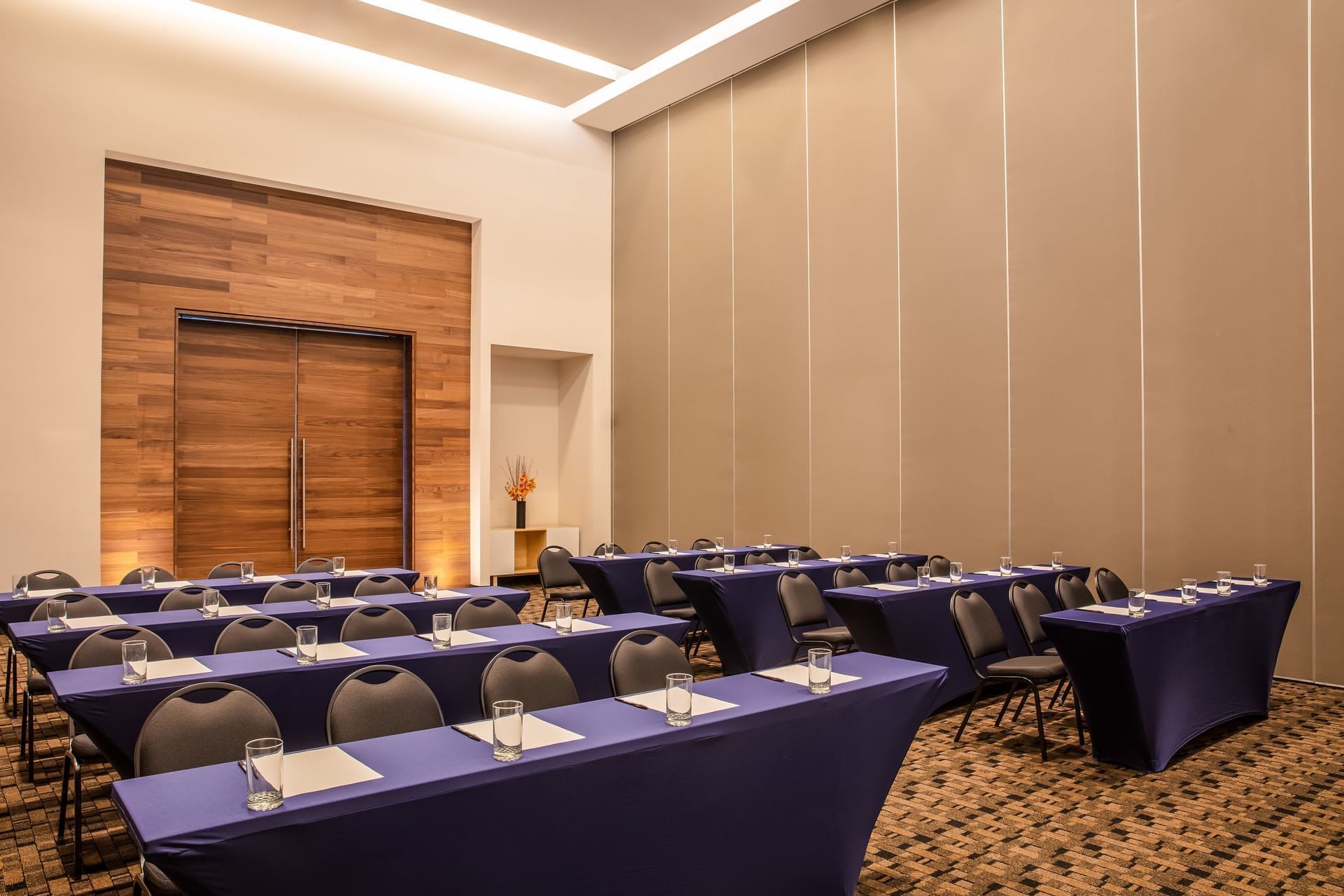 A large, modern **meeting room** at Fiesta Inn Tuxtla Gutiérrez set up in a **classroom style** configuration, perfect for **MICE** events. The room features rows of tables draped in royal blue linen, each with notepads, pens, and glasses, ready for **business delegates**. The space has high ceilings, wooden paneling by the entrance, and neutral-toned wall coverings, highlighting a professional and customizable environment for **conventions**, **workshops**, **corporate training**, and **incentive** presentations.