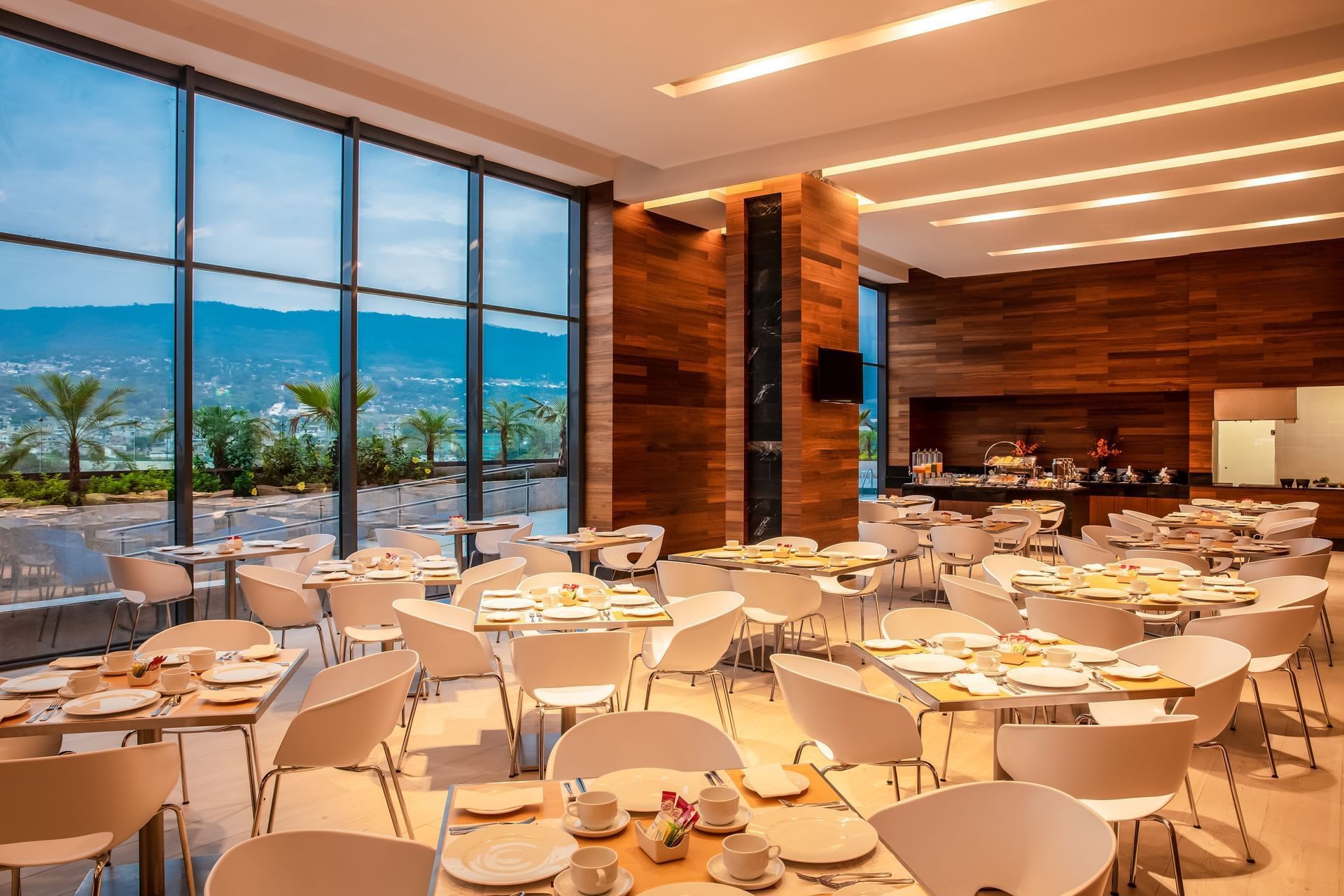 The interior of a bright, modern hotel restaurant with floor-to-ceiling windows offering a panoramic view of lush green hills and palm trees. The dining area is set with dozens of white tables and chairs, arranged for **group breakfasts** and **executive meals**. The decor features warm, wooden wall paneling, ideal for a comfortable and professional setting for **MICE** attendees, **business delegates**, and **convention** participants. It suggests high-quality **catering services** for **corporate events**