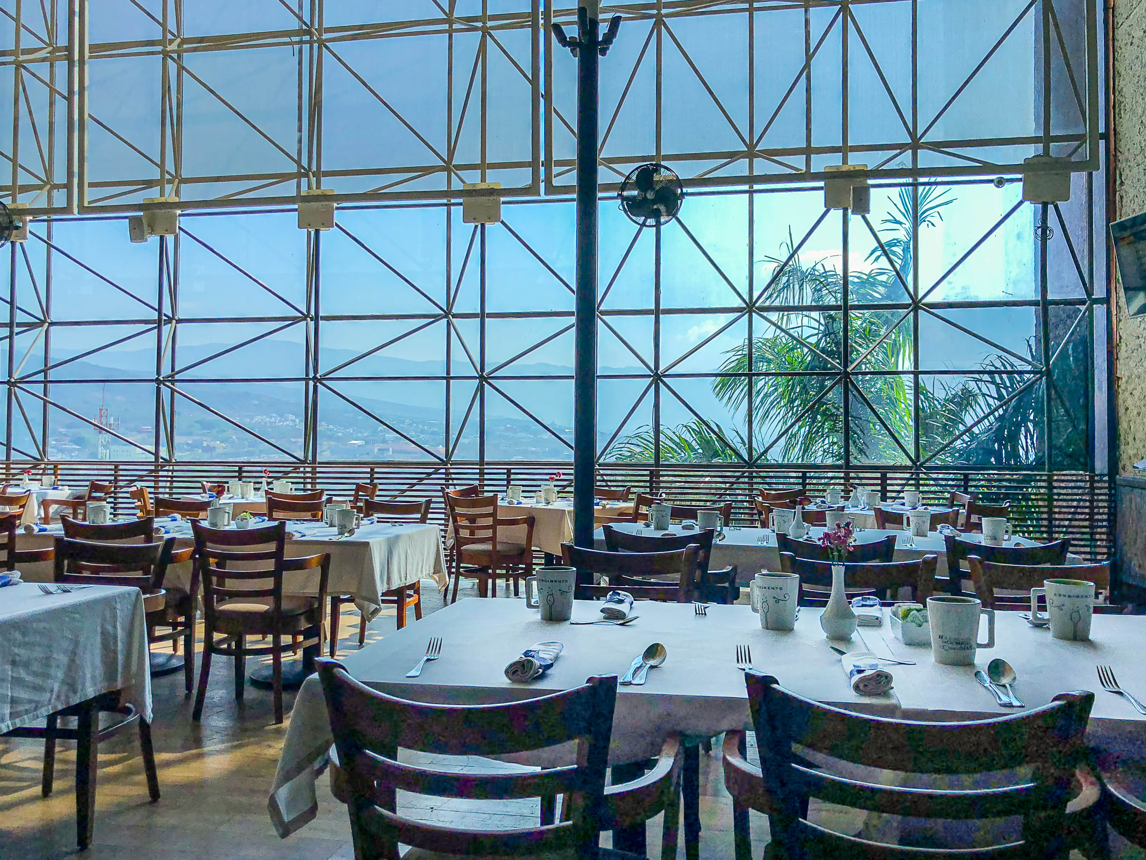 The bright dining area of the Marriott Tuxtla Gutiérrez, set up with multiple tables for a breakfast or group dining function. The room features a dramatic, floor-to-ceiling glass wall with metal framing that offers a panoramic view of the mountains and city skyline. This distinctive setting is perfect for memorable delegate meals, conference lunches, or exclusive incentive group dining experiences.