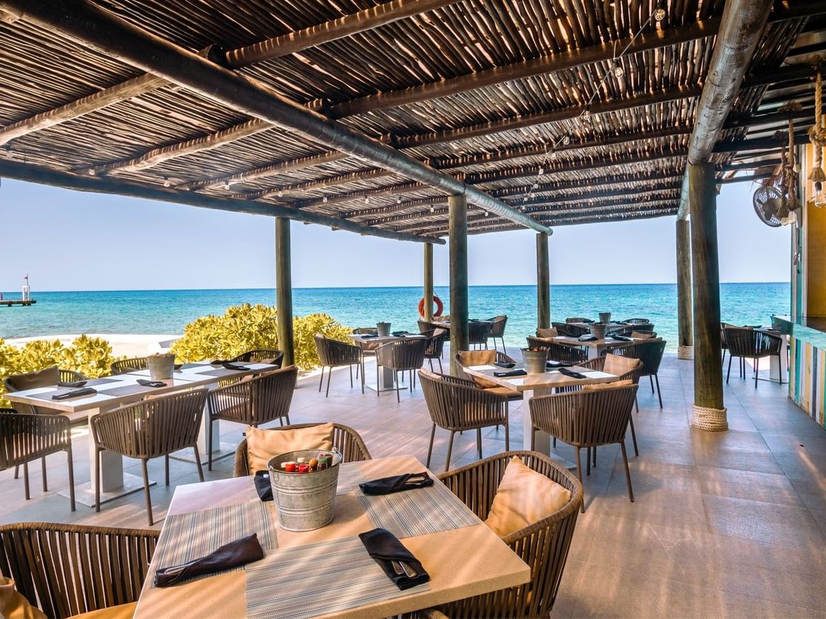 A bright, open-air restaurant at the Fiesta Americana Cozumel, featuring a rustic thatched palapa roof supported by wooden posts and modern wicker seating set on a tiled patio. The restaurant opens directly onto a sandy beach with the clear Caribbean Sea visible in the background. This attractive, casual beachfront setting is ideal for MICE group luncheons, networking receptions, and high-quality corporate hospitality events.