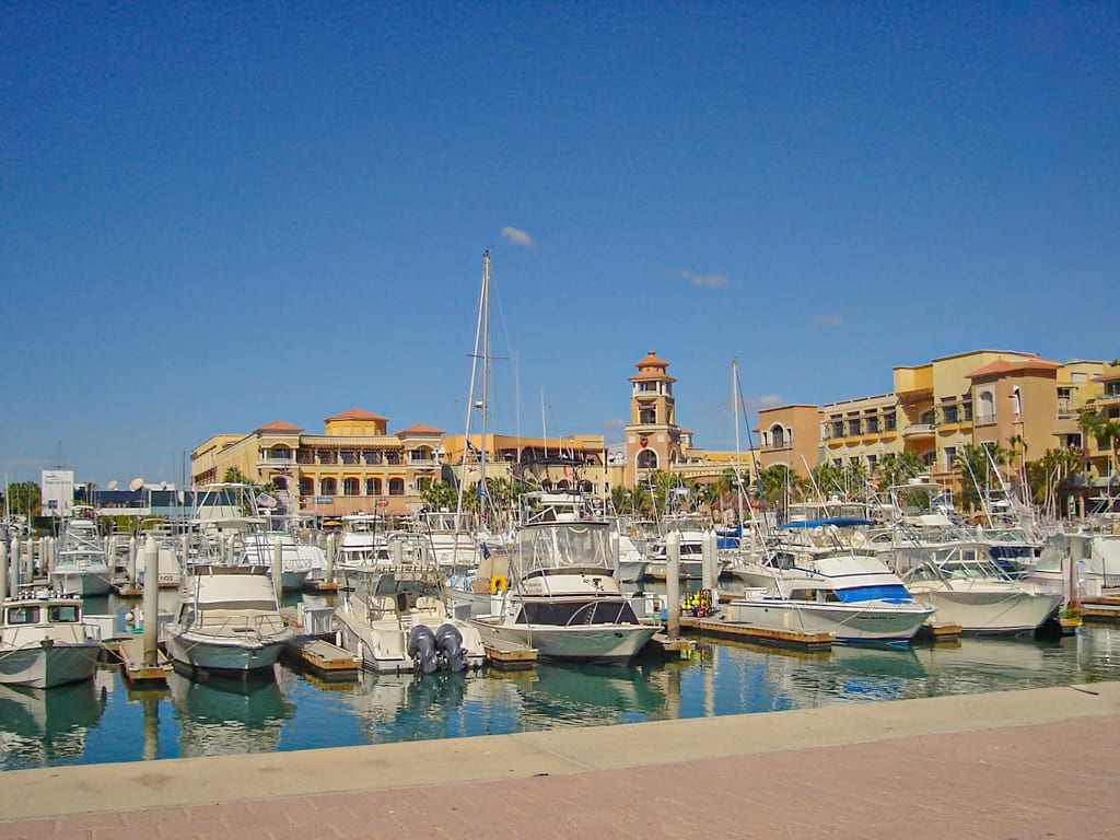 The vibrant marina in Cabo San Lucas, filled with luxury yachts and surrounded by resort-style buildings under a clear blue sky. The marina is a central hub for dining, shopping, and tours, making it a key attraction for corporate retreats and MICE groups.