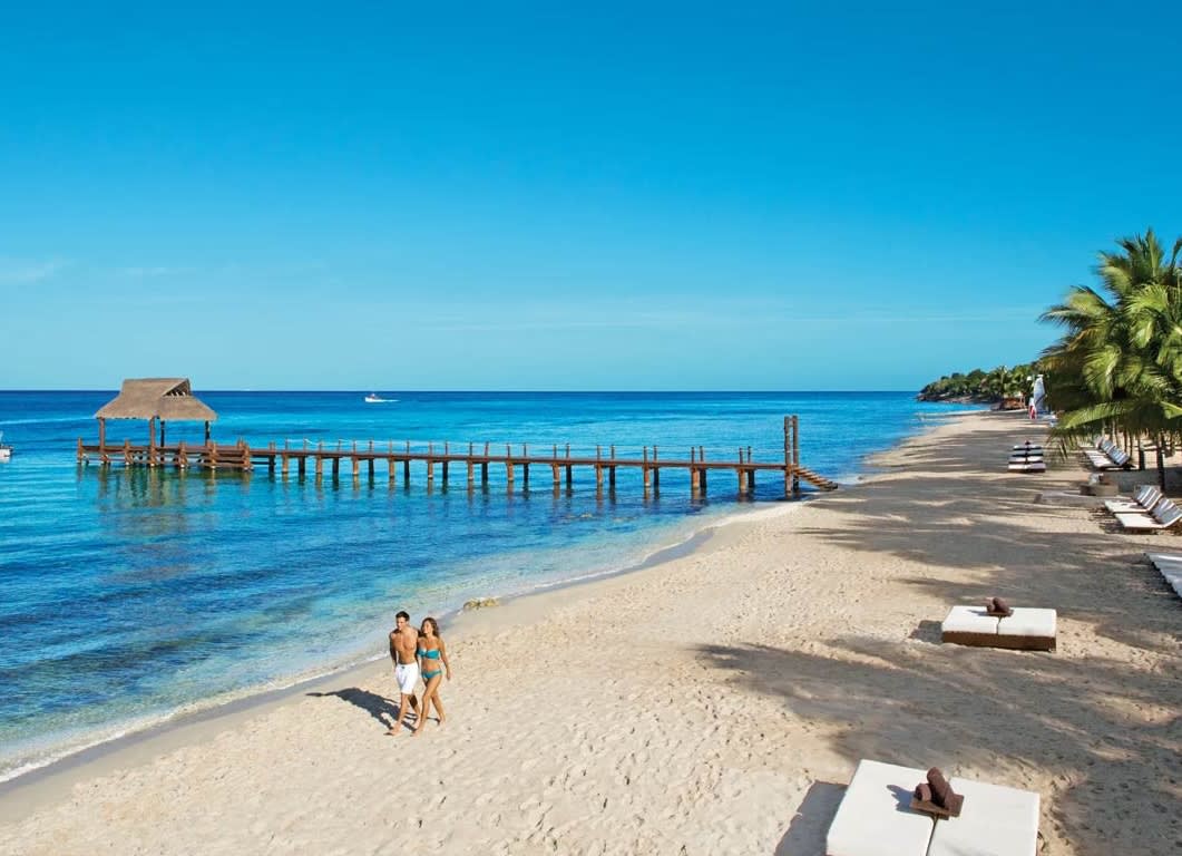 A pristine, wide, white-sand beach at Secrets Aura Cozumel, featuring a long wooden pier with a thatched-roof palapa extending over the clear turquoise Caribbean Sea. A couple is seen strolling on the beach. This idyllic, adult-only setting emphasizes luxury leisure and is perfect for incentive travel groups, providing a beautiful, natural backdrop for delegate downtime, outdoor events, and high-end corporate rewards.