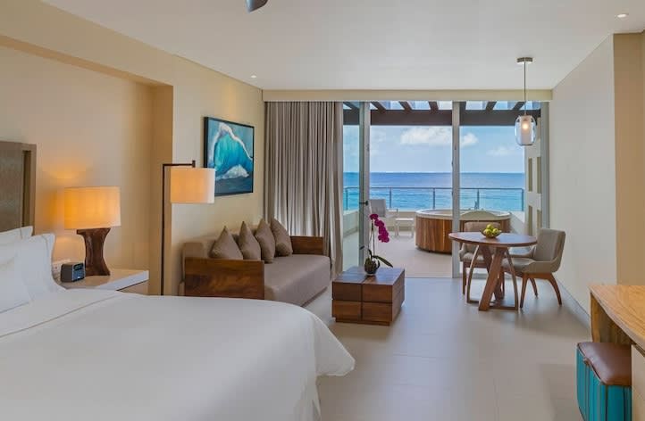 A luxurious, spacious suite at The Westin Cozumel, featuring a king-sized bed, a comfortable sofa seating area, and a dining table. Large sliding glass doors open onto a private balcony with an outdoor jacuzzi and stunning, unobstructed views of the Caribbean Sea. This premium accommodation is an exceptional reward for VIP MICE delegates and incentive travel winners, offering unparalleled relaxation and luxury after corporate events.