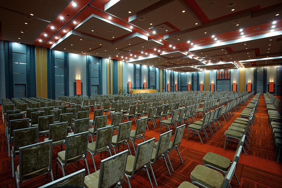 A wide-angle view of the large Grand Ballroom at the Hotel Riu Plaza Guadalajara, set up in a high-capacity theater style with hundreds of chairs facing a stage area. The room features a modern, professional design with dramatic overhead lighting and colorful accent panels. This demonstrates a flexible and expansive convention venue capable of hosting major conferences, keynote sessions, and large-scale MICE events.