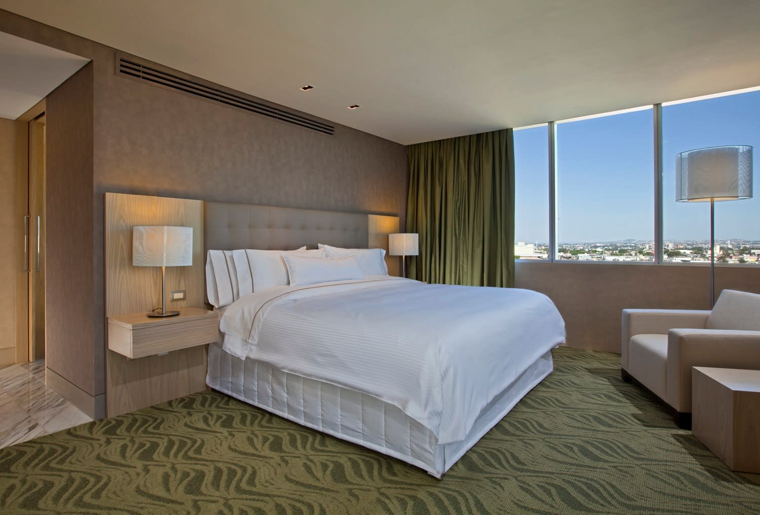 A bright, modern guest room at The Westin Guadalajara featuring a king-sized bed with crisp white linens, a neutral color palette, and a dedicated sitting area by a large window overlooking the city. The room is designed for comfort and functionality, providing ideal accommodations for business travelers, VIP speakers, and attending MICE delegates who require a productive and restful environment.