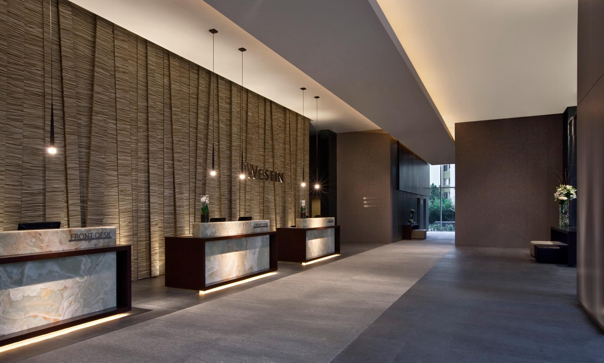 The elegant, modern reception area of The Westin Guadalajara, featuring two prominent front desks with marble tops and underlighting. The walls are clad in a textured, woven material, creating a sophisticated and welcoming environment for MICE delegates and business travelers. This space is perfectly suited for efficient attendee check-in and provides a high-end first impression for any conference or corporate event.