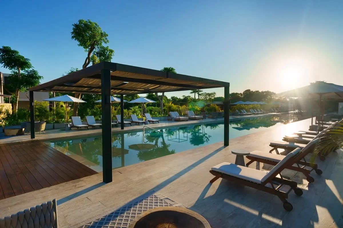 Luxury MICE poolside reception venue in Mérida: Wide, sunlit view of the Hacienda Xcanatun's large, modern swimming pool with a covered pergola area and rows of white lounge chairs. An ideal outdoor space for incentive group relaxation, networking cocktails, and high-end corporate daytime events.