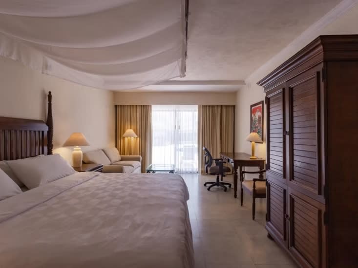 Comfortable accommodations for MICE travelers at Holiday Inn Mérida: Interior view of a spacious guest room featuring a king-size bed, a separate lounge area, a dedicated desk and chair for business work, and large windows providing a comfortable stay for convention delegates.