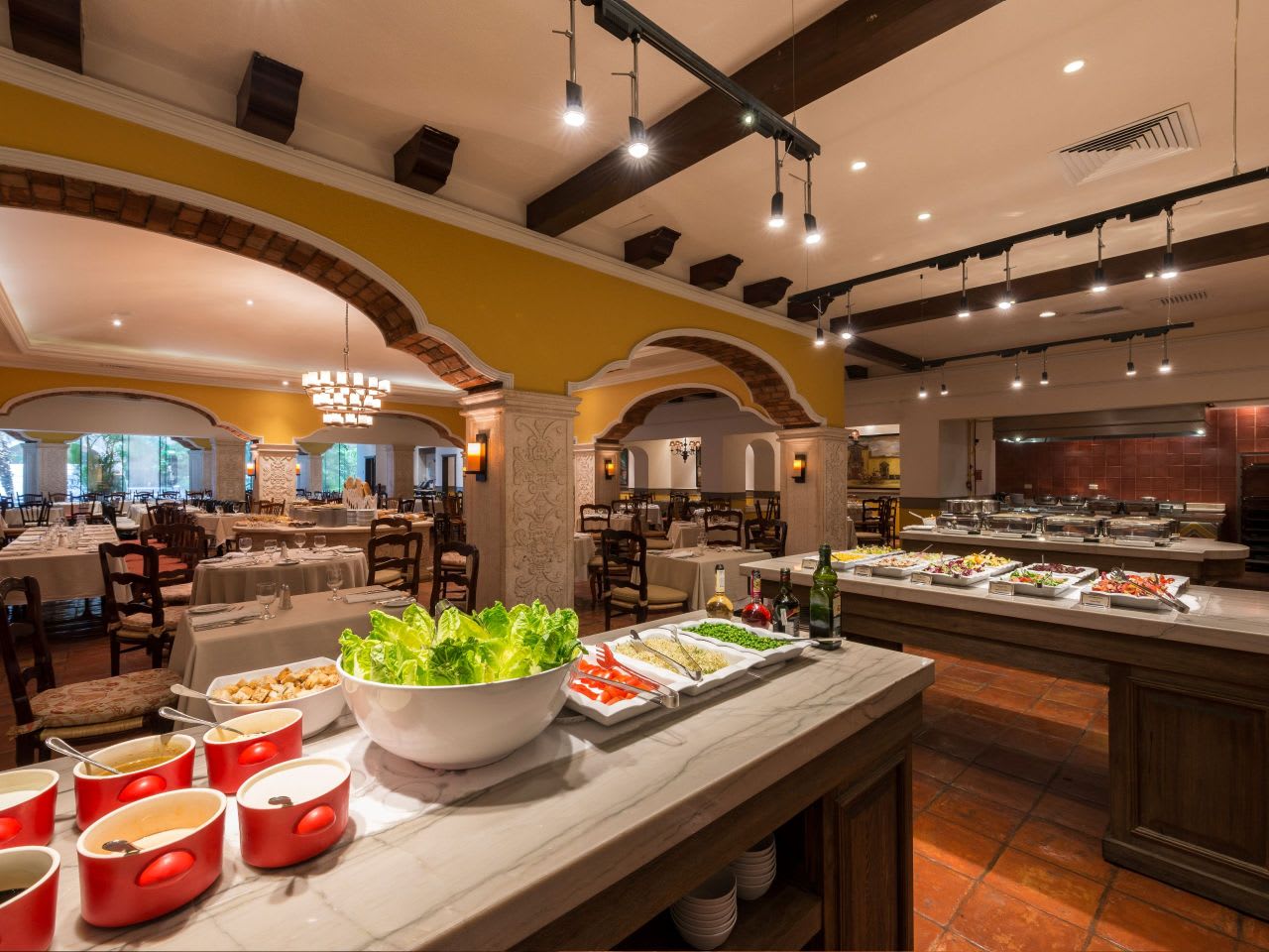 Group dining and MICE catering venue at Hyatt Regency Mérida: Interior view of The Peregrina Bistro restaurant, featuring a buffet station with fresh salad ingredients and hot food. The dining area in the background is set with tables, demonstrating a suitable space for conference lunches and delegate group catering.