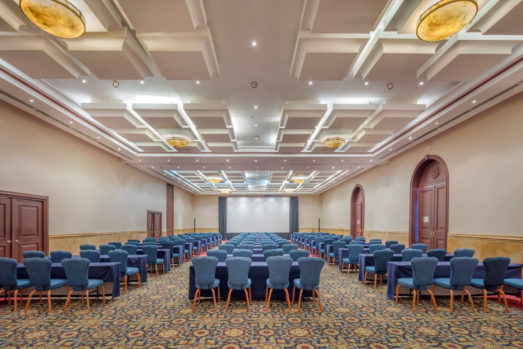 Meeting room setup for mid-sized MICE conference at Presidente InterContinental Mérida: Wide view of a large, elegant function room configured in a theater-style setup with rows of blue chairs and tables facing a projection screen. The room features decorative ceiling lights and arched doorways, ideal for corporate training and seminars.