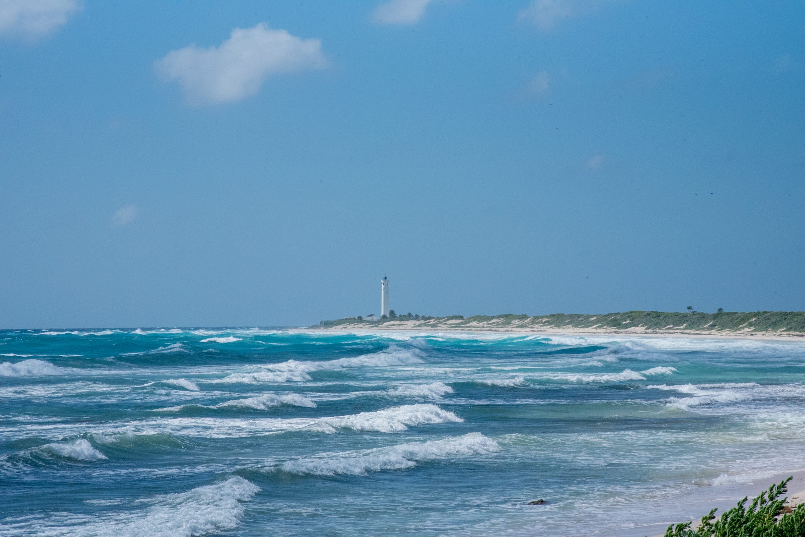 A wide-angle shot showcasing a pristine turquoise Caribbean sea with gentle waves, breaking onto a white sand beach lined with lush green vegetation. In the background, a distinct white lighthouse stands tall on a distant headland under a bright blue sky with scattered clouds. This idyllic scene highlights the natural beauty and serene environment making Cozumel a per perfect destination for  MICE events, corporate retreats, incentives, and conferences.