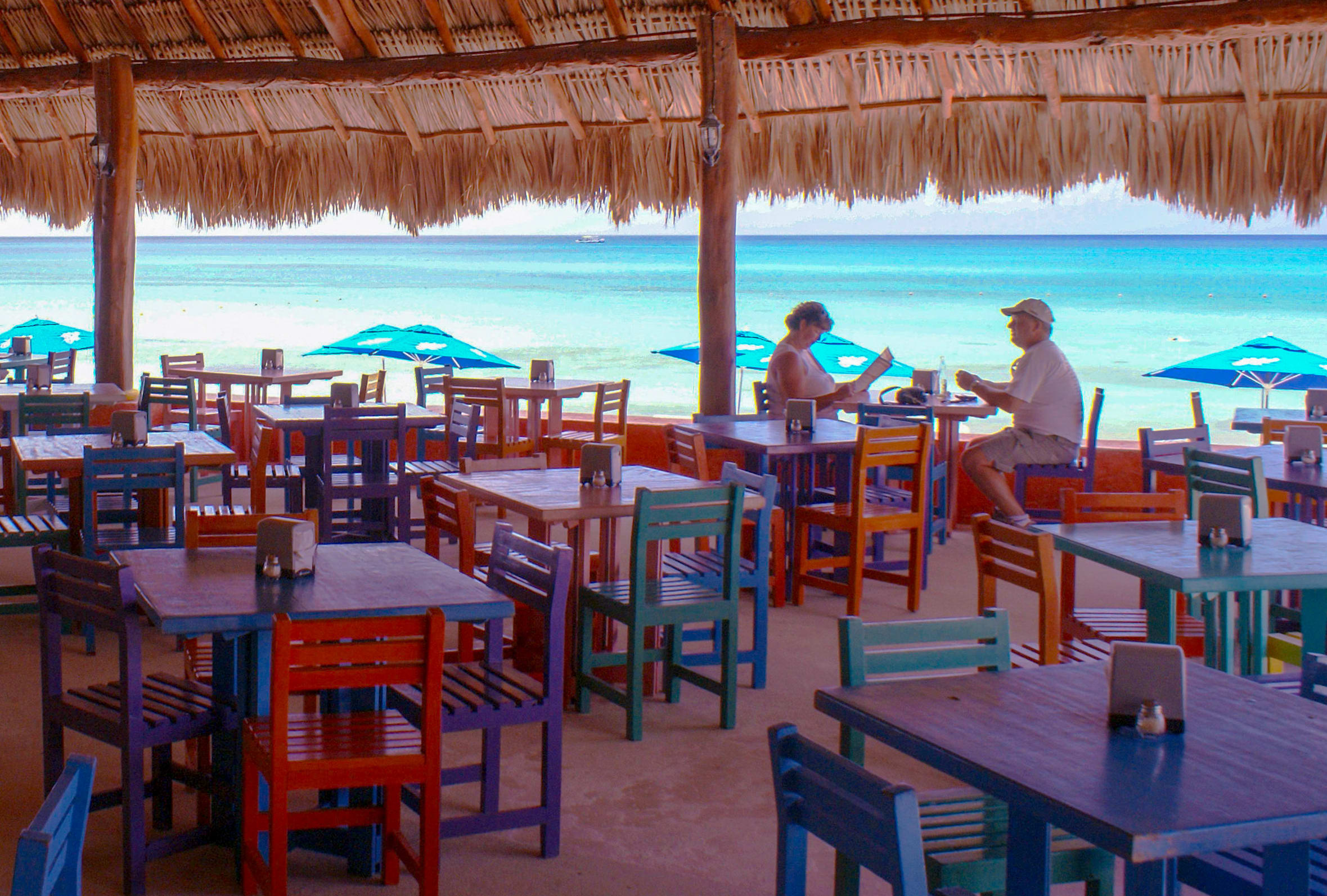 Expansive open-air beachfront restaurant setup ideal for MICE meetings, incentive travel programs, corporate conferences, and exhibitions in a luxurious Caribbean tropical resort: thatched palapa roof with wooden beams and hanging lantern over colorful wooden tables and chairs in vibrant reds, blues, greens, and purples; scattered salt and pepper shakers on surfaces; in the foreground, a relaxed couple – a woman in white tank top seated reading a tablet or book, and a man in white shirt and cap lounging nearby; stunning turquoise ocean backdrop with white sand beach, scattered blue beach umbrellas, gentle waves, distant boat on deep blue sea under clear sky – perfect seaside venue for team-building events, networking lunches, and high-end incentive rewards in paradise