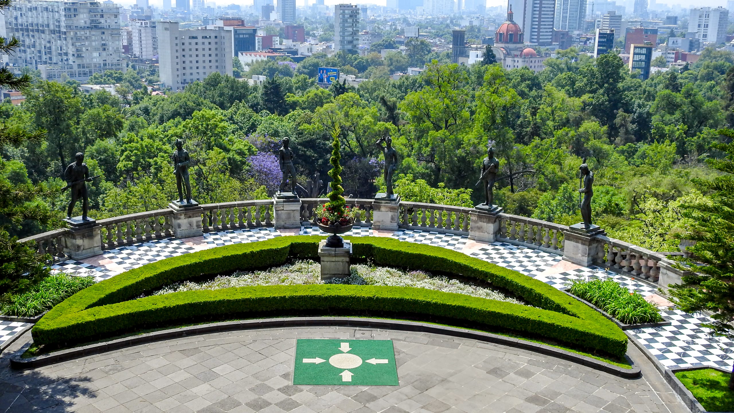 A panoramic view from Chapultepec Castle in Mexico City, overlooking a lush green park and the city skyline. The foreground features a manicured terrace with classical statues and a checkered tile floor. This breathtaking view is an inspiring backdrop for MICE industry events and incentive travel.