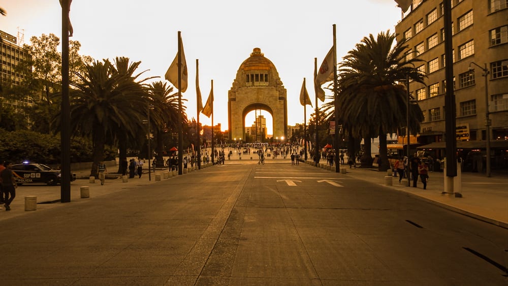 A golden-hour view of the Monument to the Revolution in Mexico City, with a wide pedestrian walkway leading to the archway. The scene is lined with palm trees and streetlights and is busy with people. This landmark is a prominent venue for a souvenir photo for MICE industry events and incentive travel.