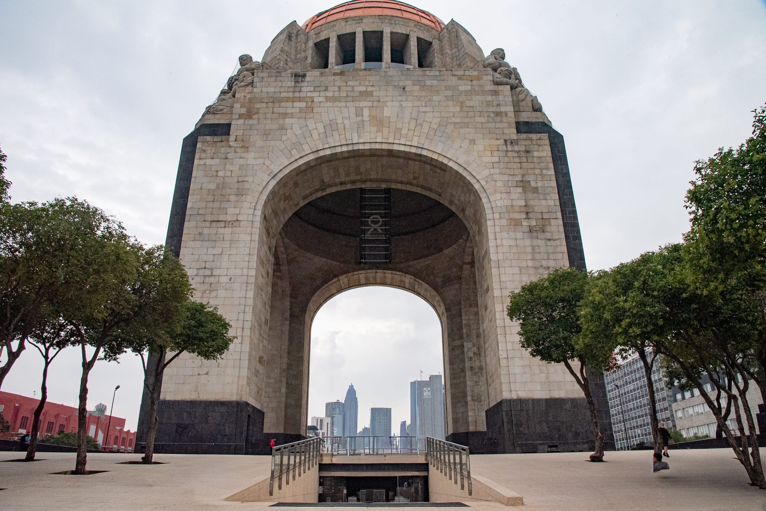 The Monument to the Revolution in Mexico City, a large, stone archway with a bronze dome, viewed from a pedestrian plaza. Modern skyscrapers are visible through the arch. This iconic landmark is a prime location for MICE industry events and incentive travel.