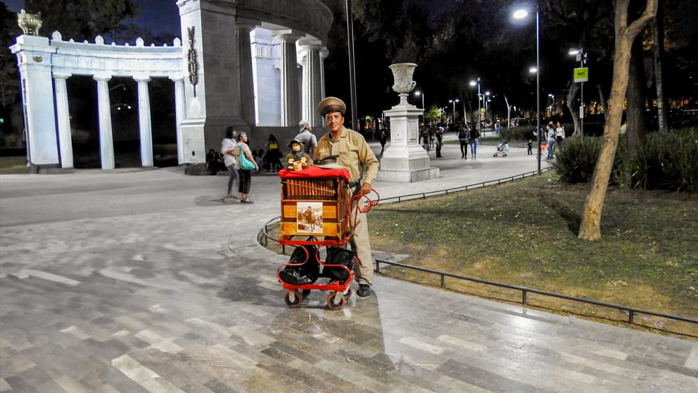 A traditional Mexican organ grinder, known as a 'cilindrero,' stands with his instrument on a street at night in Mexico City. The background shows a public park with people walking and a historic stone archway. This image captures the unique cultural experiences available in Mexico City, perfect for MICE industry event entertainment and incentive travel.