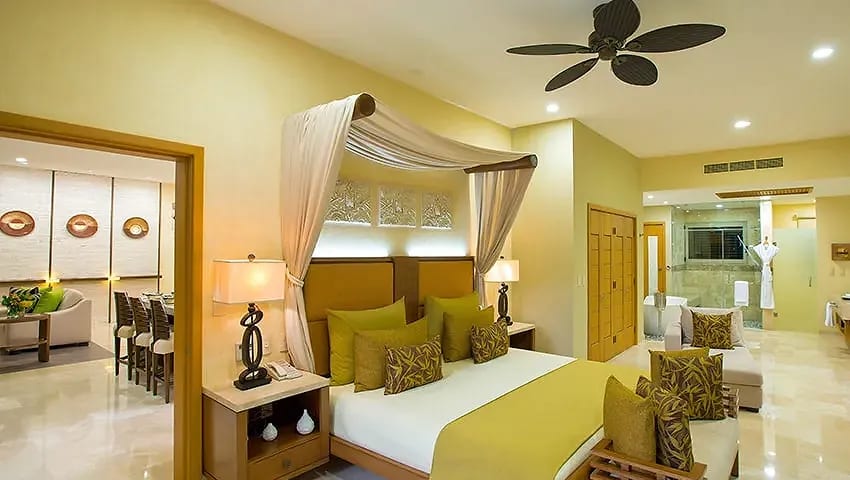 Luxurious bedroom suite at Garza Blanca Preserve Resort & Spa, a top MICE venue in Mexico for meetings, incentives, conventions, and exhibitions, featuring a canopy bed with green and yellow accents, ceiling fan, bedside lamps, tropical decor, adjoining dining area with table and chairs, and visible spa-like bathroom ideal for attendee comfort during incentive travel and corporate events in Puerto Vallarta.