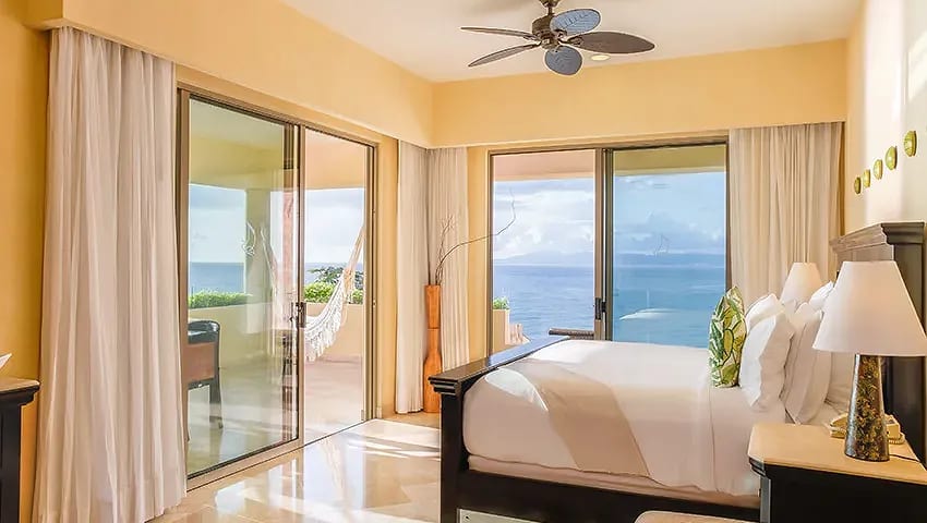 Luxurious ocean-view bedroom at Garza Blanca Preserve Resort & Spa, a premier MICE venue in Mexico for meetings, incentives, conventions, and exhibitions, featuring a king-size bed with white linens and green accents, ceiling fan, sheer curtains, bedside lamps, and balcony with hammock overlooking the Pacific Ocean and tropical landscape ideal for attendee relaxation during incentive travel programs in Puerto Vallarta.