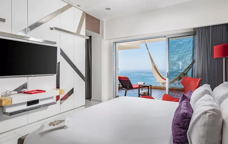 Modern ocean-view bedroom at Hotel Mousai Puerto Vallarta, an adults-only luxury MICE venue in Mexico for meetings, incentives, conventions, and exhibitions, featuring a white bed with purple pillows, wall-mounted TV, geometric closet design, red floor lamp, and balcony with hammock and lounge chairs overlooking Banderas Bay ideal for attendee relaxation in incentive travel programs.