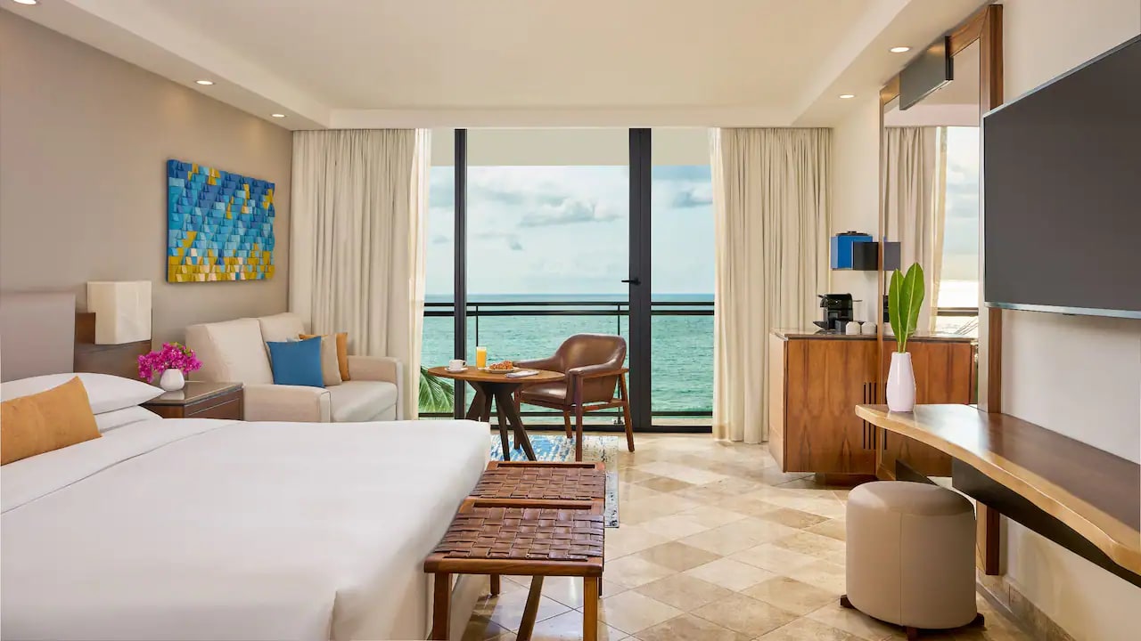 Luxurious ocean-view suite at Hyatt Ziva Puerto Vallarta, a premier all-inclusive MICE venue in Mexico for meetings, incentives, conventions, and exhibitions, featuring a king-size bed with orange pillow, beige sofa, wooden benches, breakfast table, wall-mounted TV, fresh flowers, and balcony overlooking Banderas Bay ideal for attendee relaxation during incentive travel programs.