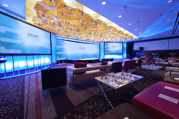 Modern, brightly lit executive meeting or lounge area for a MICE event. The room features comfortable sofas, lounge chairs, a geometric ceiling fixture, and three large screens displaying the "ENCORE" logo. Perfect for VIP receptions, networking, or executive general sessions. Keywords: Executive meeting setup, MICE lounge area, corporate reception AV, innovative event design, high-end meeting technology.