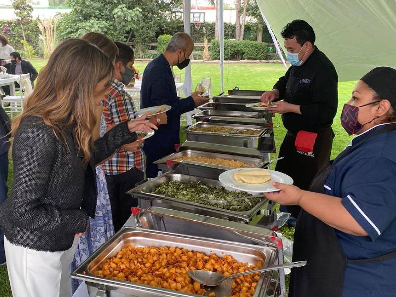 Attendees at an outdoor corporate event lining up at a self-serve buffet line. Professional caterers, wearing masks, are serving authentic Mexican dishes like potatoes and greens. This image captures a casual but professional business lunch, conference reception, or team incentive event setup with outdoor catering services.