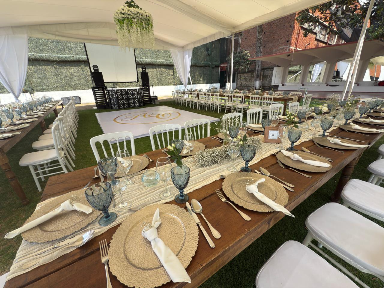 A long, elegantly set outdoor dining table for a large corporate event, wedding, or gala. The rustic wooden table is set with blue glassware, woven placemats, white napkins, and professional flatware. The event venue features a white tent, a dance floor, and a projector screen, ideal for a memorable MICE industry incentive dinner or corporate gala.