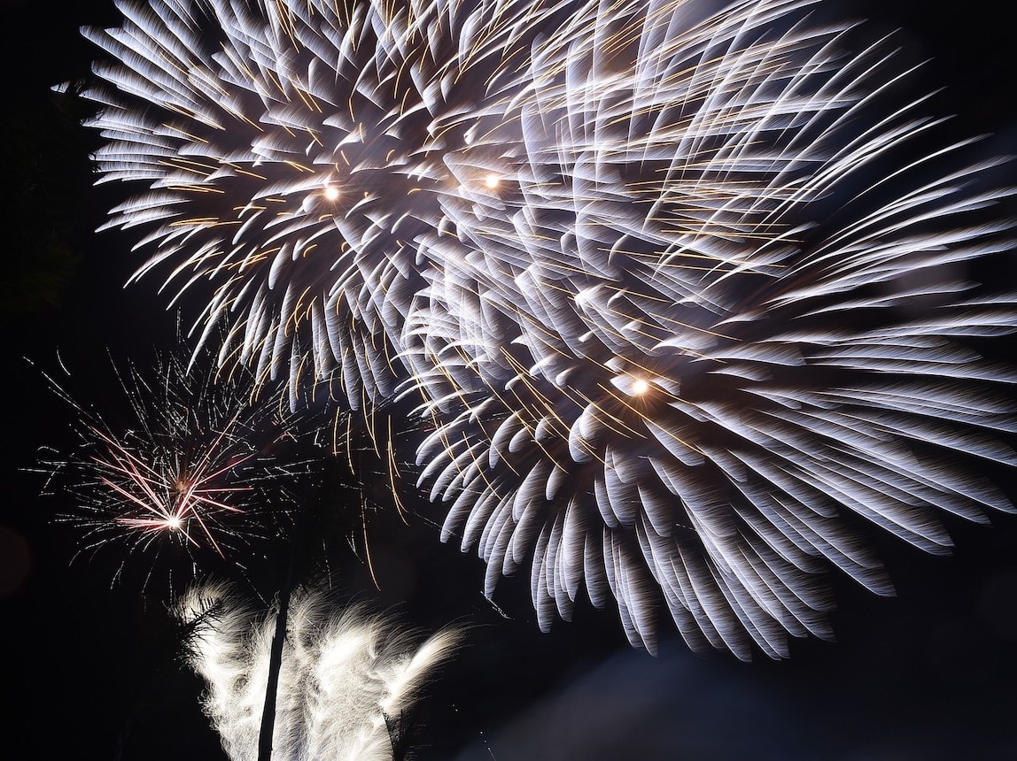 A night-time outdoor event scene on a beach or deck with red and gold fireworks exploding in the sky above. Guests are taking photos and watching the show near a draped, illuminated stage. An excellent backdrop for an incentive travel event, corporate gala dinner, or MICE reception in a tropical venue.