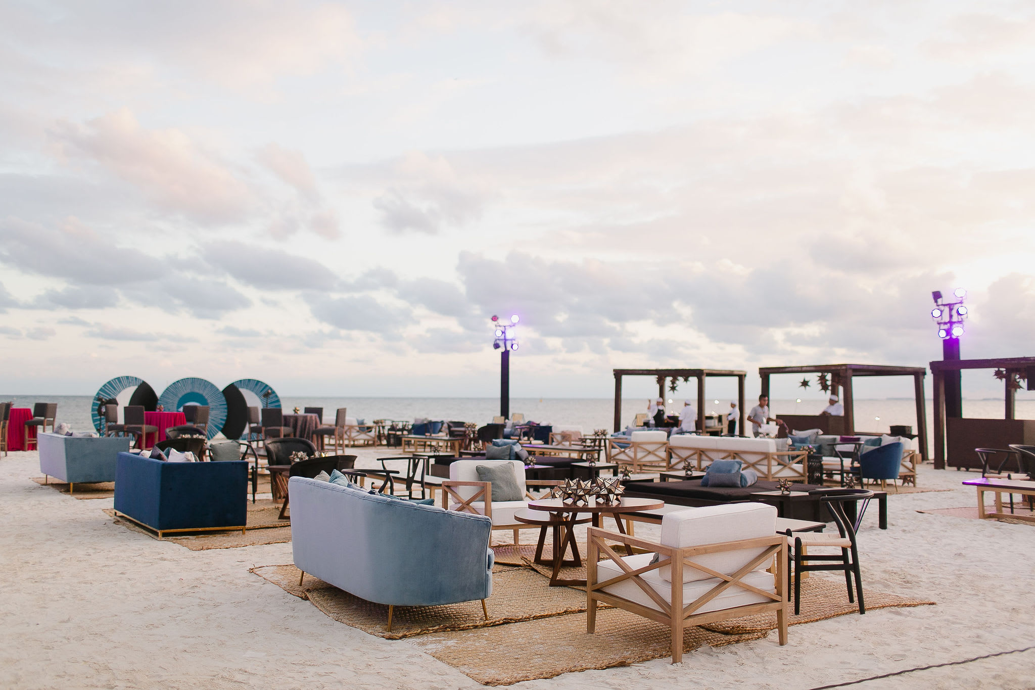 A comfortable and stylish beach lounge setup featuring modern sofas and armchairs in muted blues and whites, placed on woven rugs on the sand. The sunset glow provides lighting, perfect for a casual cocktail reception, networking event, or post-conference relaxation area for MICE attendees in Mexico.