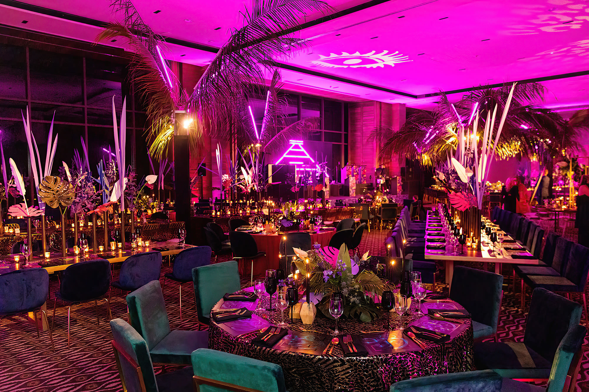 A large indoor event space transformed into a lavish, jungle-themed corporate gala dinner. The room is dramatically lit with pink and purple neon lights, featuring palm trees, velvet emerald chairs, and geometrically patterned tablecloths. Ideal for a high-impact incentive event, conference after-party, or themed MICE function.