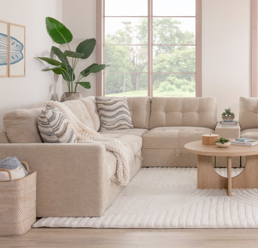 A beige modular sectional sofa with decorative pillows sits in a bright room with large windows.