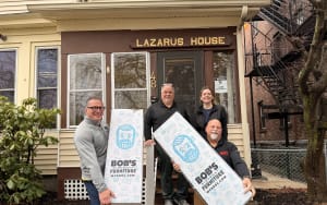 Four people holding large white boxes with blue graphics and text "BOB'S DISCOUNT FURNITURE MYBOBS.COM" stand in front of a building with a sign "LAZARUS HOUSE" above the door, indicating a furniture donation or delivery.
