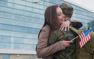 A woman warmly hugging a soldier in camouflage uniform holding a small American flag, outside a modern glass building.