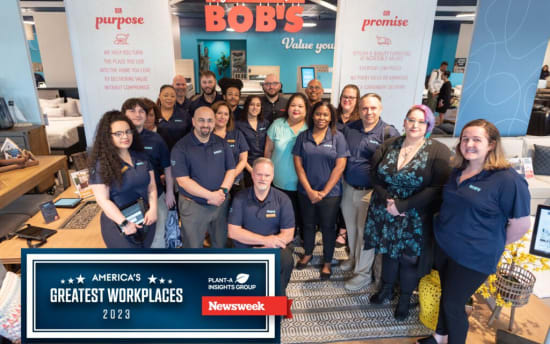 Team of employees in navy blue BOBS uniforms standing inside a modern furniture store with signs reading "Welcome to BOBS," "Our purpose," and "Our promise." Text includes: "America’s Greatest Workplaces 2023," "Plant-A Insights Group," and "Newsweek." The store features stylish, light wood furniture and home decor.