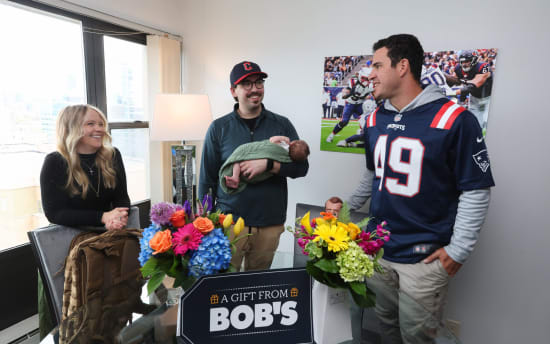 Three adults stand around a glass table with two vibrant floral arrangements in blue, orange, yellow, and pink hues. One person holds a baby wrapped in a green blanket. A sign on the table reads "A GIFT FROM BOB'S." The setting is a bright room with large windows and a sports-themed wall art featuring a football game. One adult wears a navy Patriots jersey with number 49.