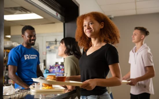 Young people standing in line at a cafeteria counter holding trays with food, including sandwiches and drinks, in a casual indoor setting.