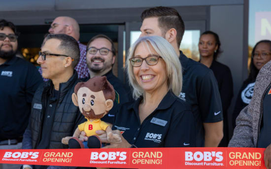 Group of people at a grand opening event holding a red ribbon with text "BOB'S Discount Furniture. GRAND OPENING!" One person holds a plush doll wearing a yellow shirt with a red "B." Several individuals wear black shirts with the Bob's Discount Furniture logo.