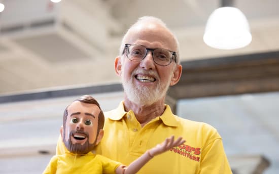 Older man wearing a yellow polo shirt with "Bob's Furniture" logo holding a yellow-shirted articulated doll in a bright indoor setting.