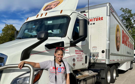 Woman in casual gray t-shirt, blue jeans, and white sneakers leaning on a white Bob’s Discount Furniture delivery truck with logo and contact details.