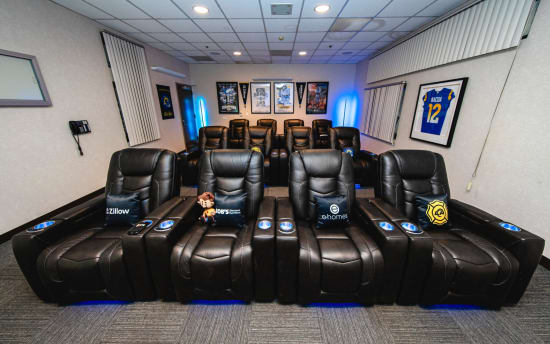 Black leather reclining theater chairs with branded pillows from Zillow, Bob's Discount Furniture, and e-homes, arranged in two rows with blue LED lighting in a modern home theater room.
