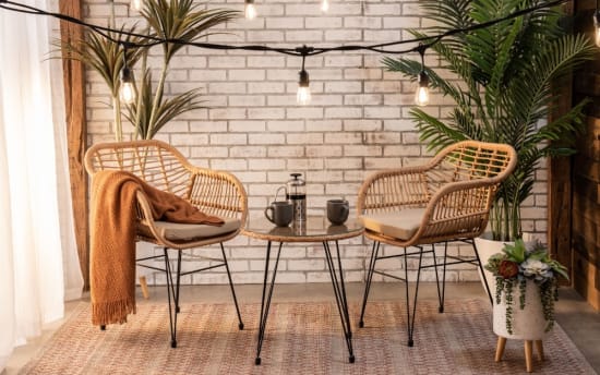 Cozy seating area with two rattan chairs featuring beige cushions, a round glass-top table, warm string lights, and green potted plants on a patterned rug.