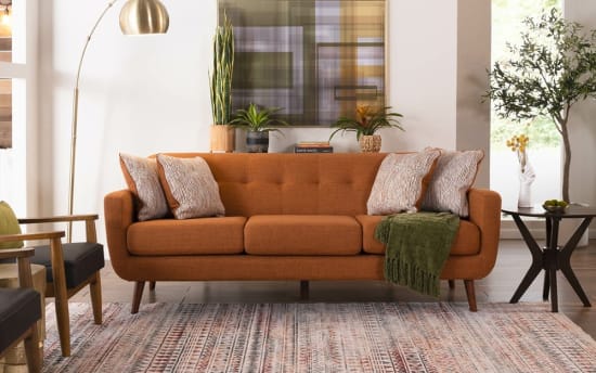 Mid-century modern burnt orange fabric sofa with tufted cushions, wooden legs, patterned throw pillows, green throw blanket, and decorative plants.