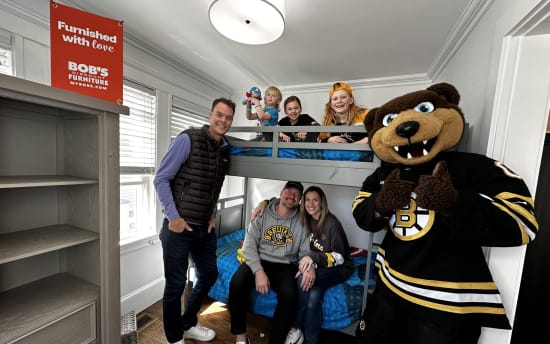 A family gathered in a bedroom with gray bunk beds and blue bedding, alongside a person in a Boston Bruins mascot costume. A red sign reads "Furnished with love BOB'S DISCOUNT FURNITURE MYBOBS.COM." The room features white walls, hardwood floors, and a gray wooden shelving unit.