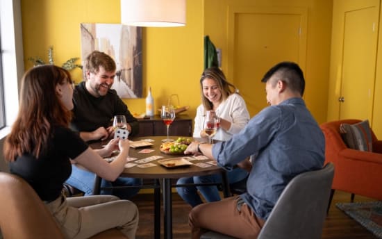 Four people sitting around a round wooden table playing cards with wine glasses and a fruit platter in a cozy yellow-walled room.