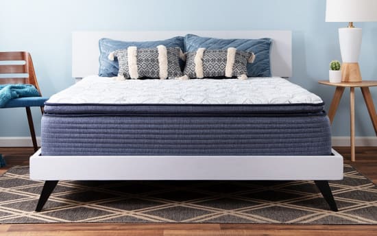 Modern bedroom setup featuring a white platform bed with a thick blue mattress, white quilted cover, decorative blue and patterned pillows, wooden side table with lamp and cactus, and a wooden chair with blue cushion against a light blue wall.